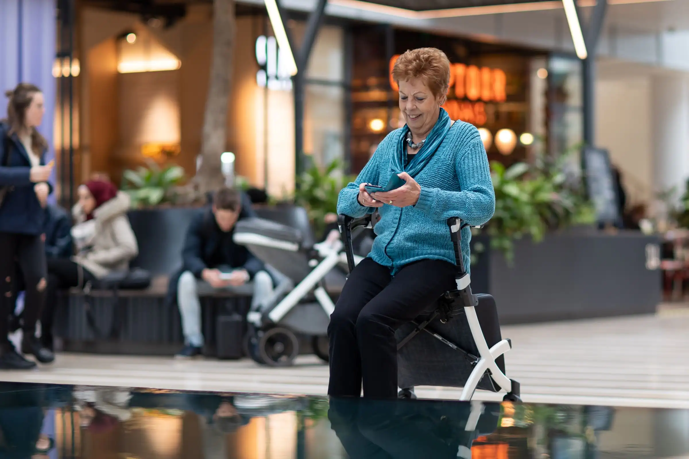 An older woman sitting on her rollator walker in a shopping centre, checking her phone