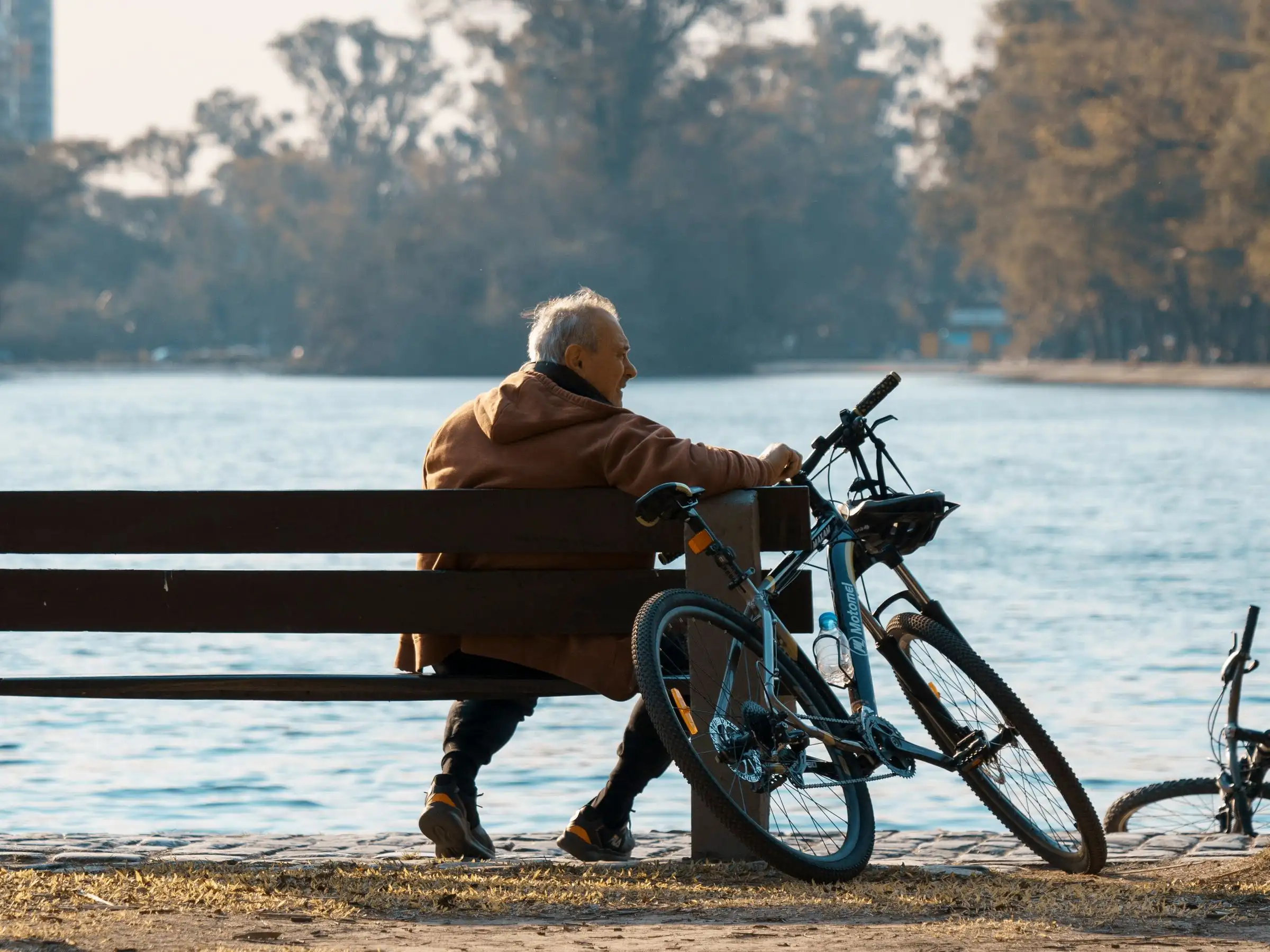 An older man resting on a bench by the water, his bike parked beside him
